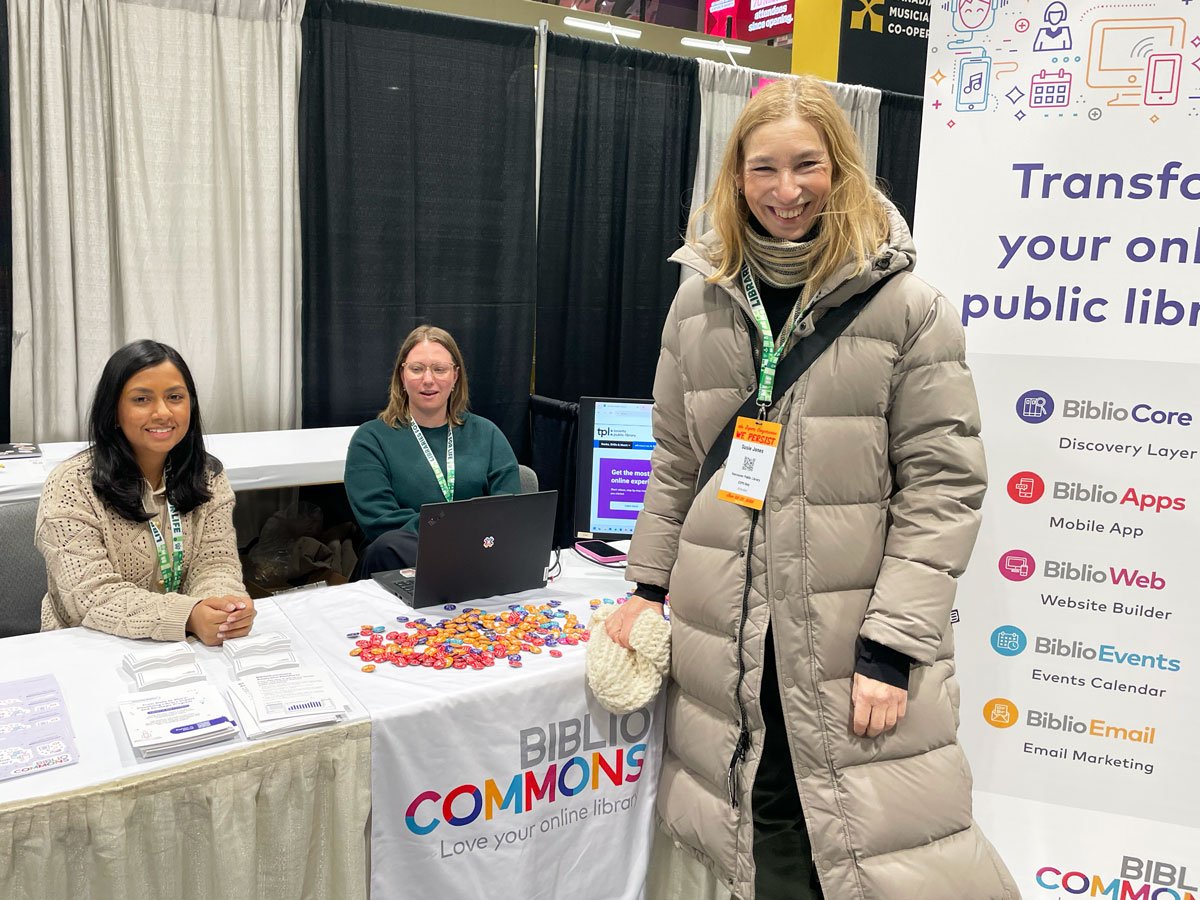 Three people stand and sit at the BiblioCommons exhibit booth, smiling and talking. The table displays brochures, buttons, and a laptop, with a banner listing BiblioCommons products behind them.